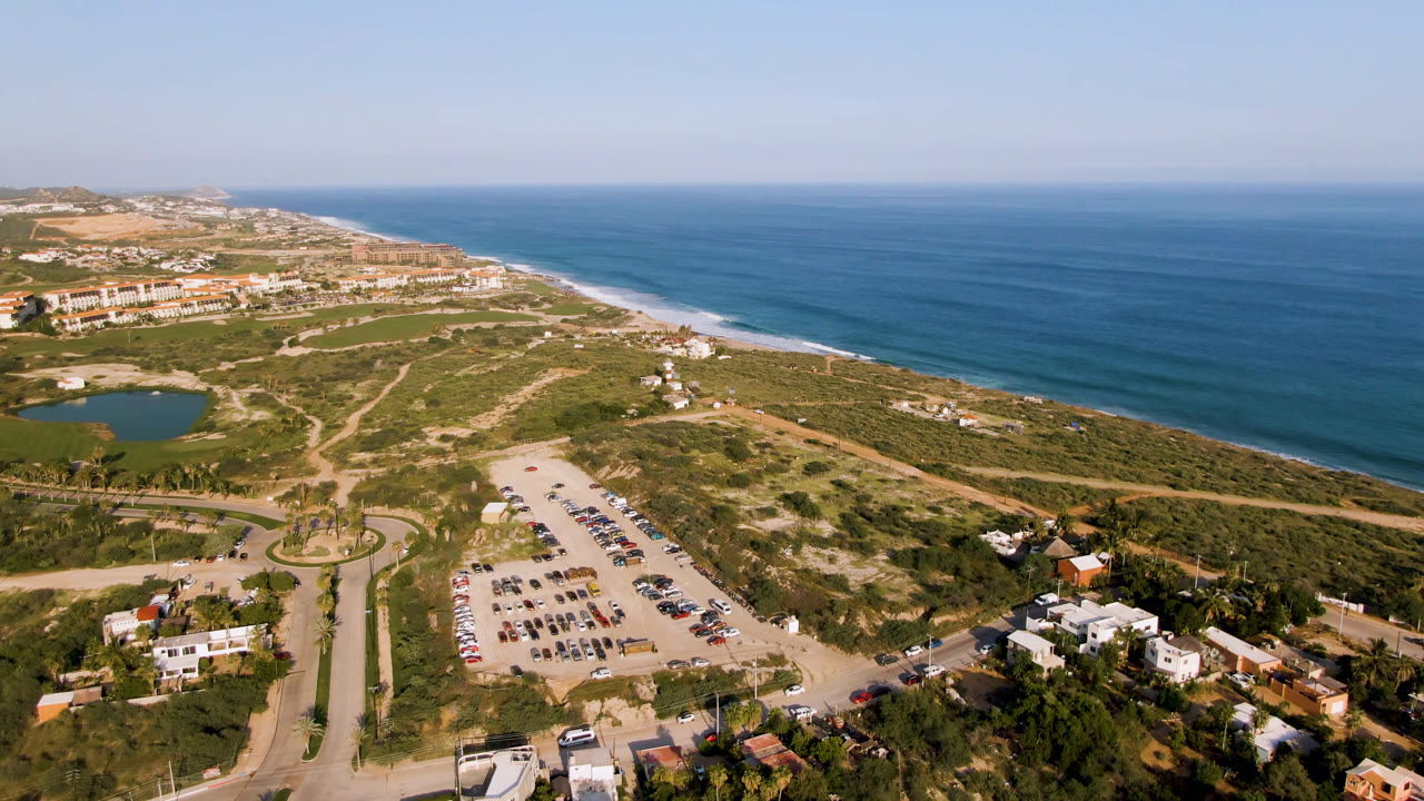 Wide drone view of blue ocean, beach, access roads, parking, and coastal buildings.