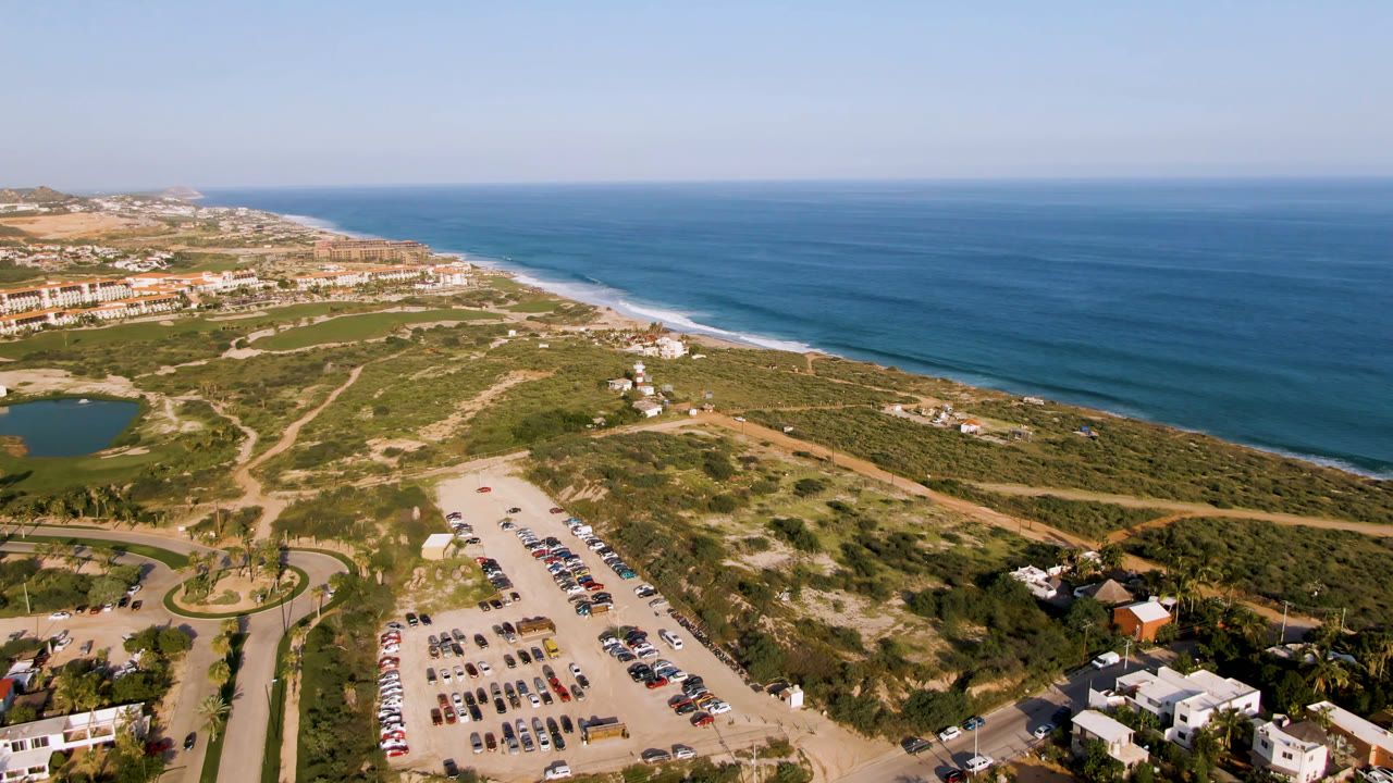 High aerial frame over Puerto Los Cabos with the parking lot lower in frame, green coastal vegetation and dirt tracks in the center, low buildings along the shore, blue ocean beyond, and no visible people.