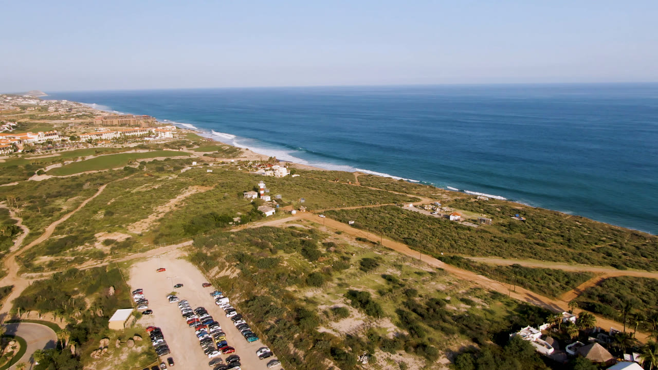 Puerto Los Cabos drone view with rows of parked cars near the lower left, tan dirt roads crossing green coastal scrub, small buildings near the beach, and the ocean horizon under clear sky, with no visible people.