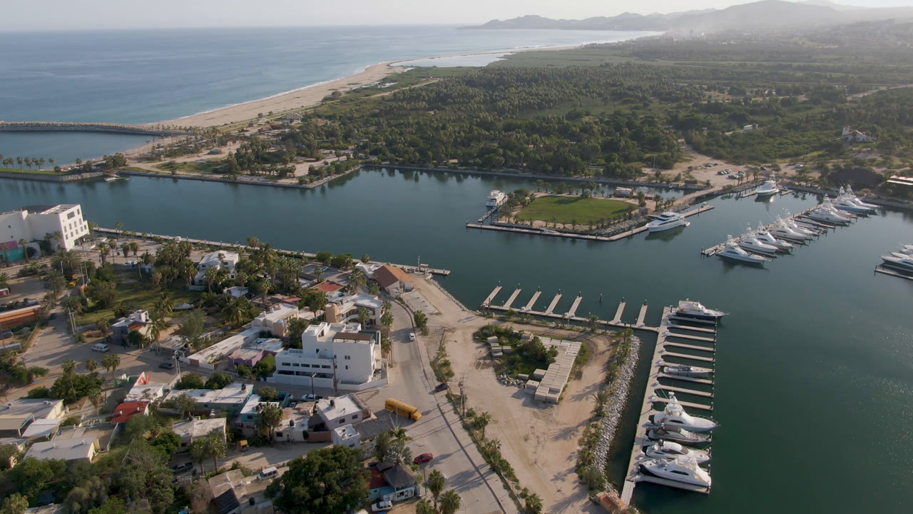 Aerial marina view with docked boats, waterfront buildings, beach, ocean, and distant hills.