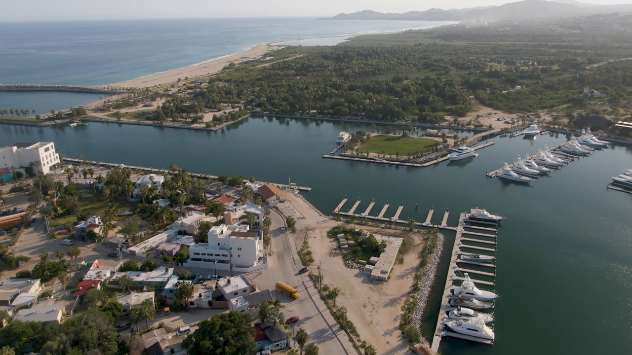 Second aerial frame over Puerto Los Cabos, Mexico, with empty marina slips in the lower right, docked yachts to the right, a green lawn near the water, beach and ocean in back, and no visible people.