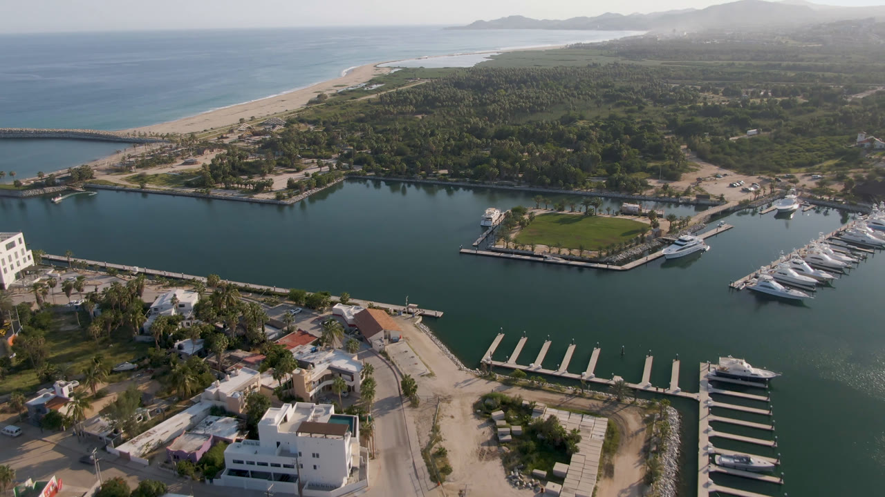 High aerial frame over Puerto Los Cabos showing marina water, a grass-covered island dock, yachts, shoreline roads, beach, ocean, distant hills, and no visible people.