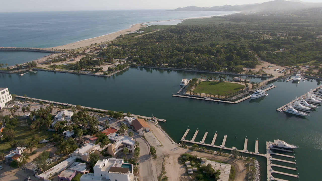A second Puerto Los Cabos aerial frame centers the marina channel, docked yachts, empty slips, palm-lined shoreline, sandy beach, ocean, and no visible people.