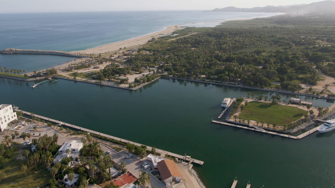 Wide aerial frame over Puerto Los Cabos showing green marina water in front, a sandy beach and blue ocean beyond, waterfront structures, palm trees, and no visible people.