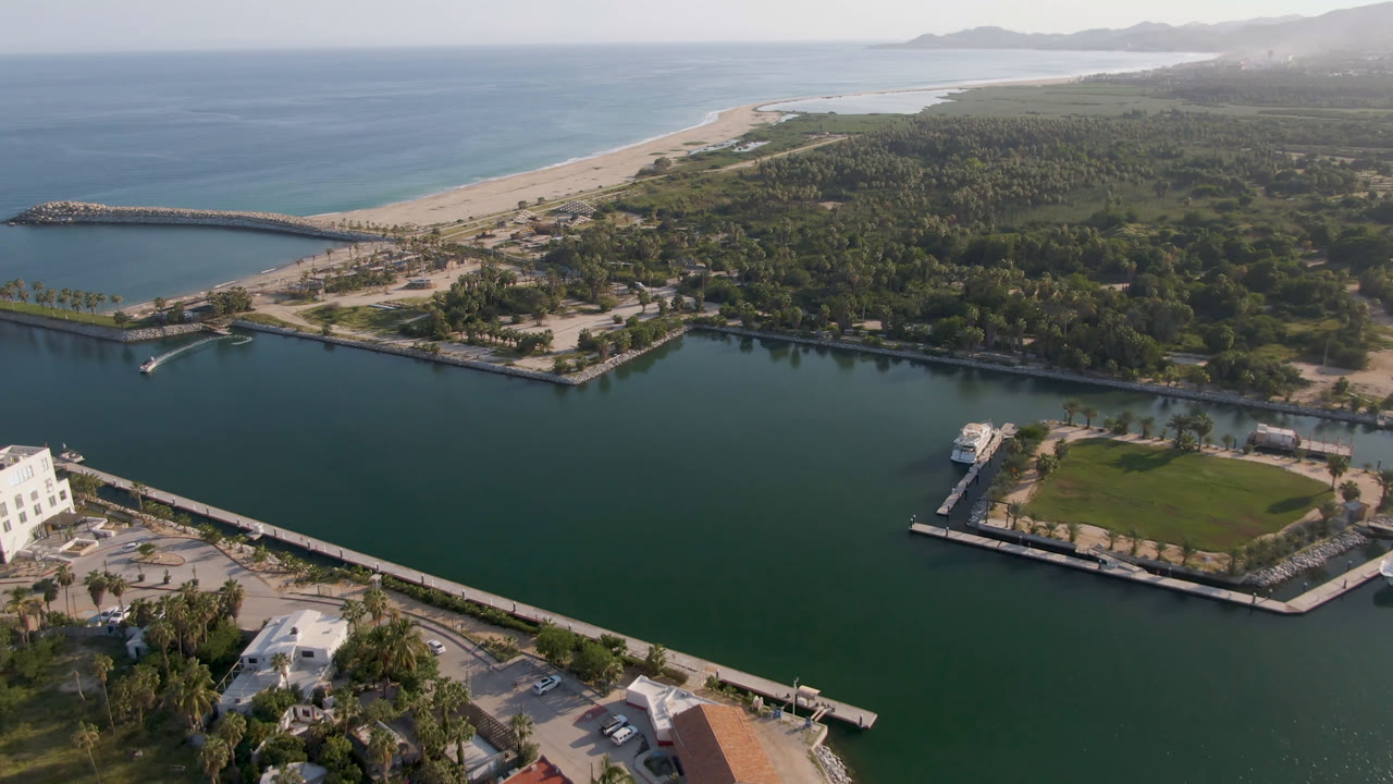 Aerial view in Puerto Los Cabos with the marina channel across the lower frame, a small boat near a dock, beach sand and ocean on the horizon, and no visible people.