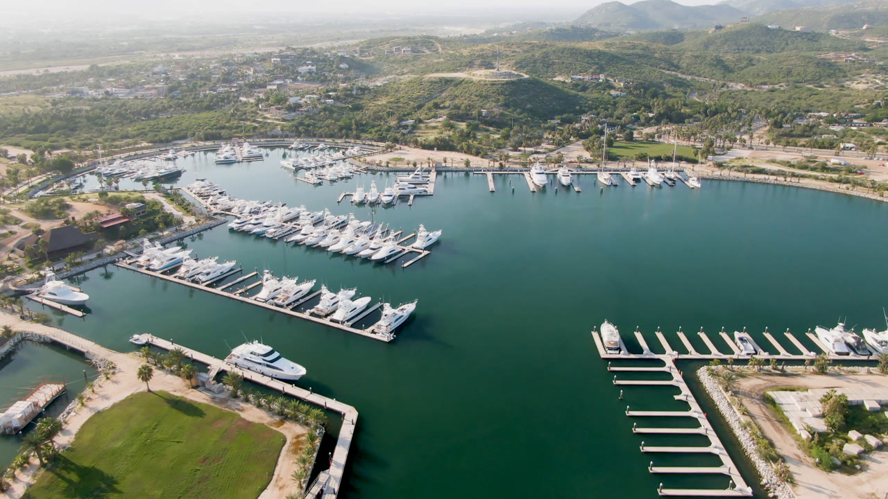 Opening aerial frame over Puerto Los Cabos marina, showing rows of white boats at docks, green water, a lawn at the lower edge, shoreline roads, hazy hills, and no visible people.
