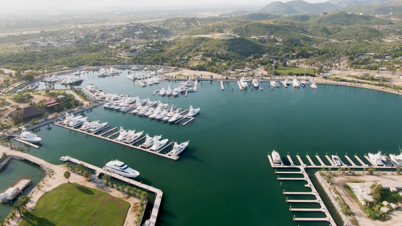 Second aerial frame of Puerto Los Cabos marina with white yachts arranged along finger docks, an open basin of green water to the right, shoreline roads, hills, and no visible people.