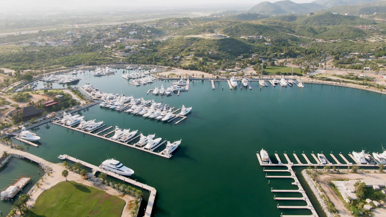 Drone view across marina water with rows of docked boats and hillside background.