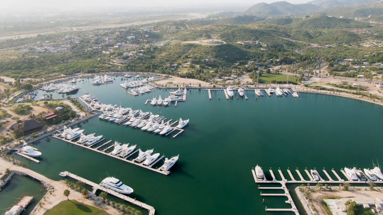 Aerial frame over Puerto Los Cabos marina showing rows of white boats tied to docks, green-blue water, shoreline roads, dry hills, and no visible people.