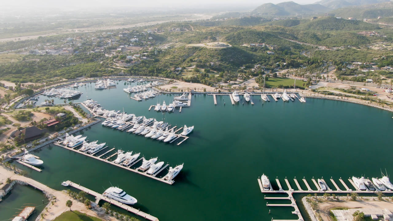 High aerial view in Puerto Los Cabos with a large marina basin, clustered docked yachts, pale roads along the shore, hazy hills, and no visible people.