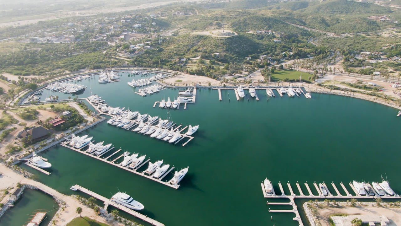 Opening aerial frame over Puerto Los Cabos, Mexico, showing rows of white yachts at floating docks, green-blue marina water, roads, low buildings, and no visible people.