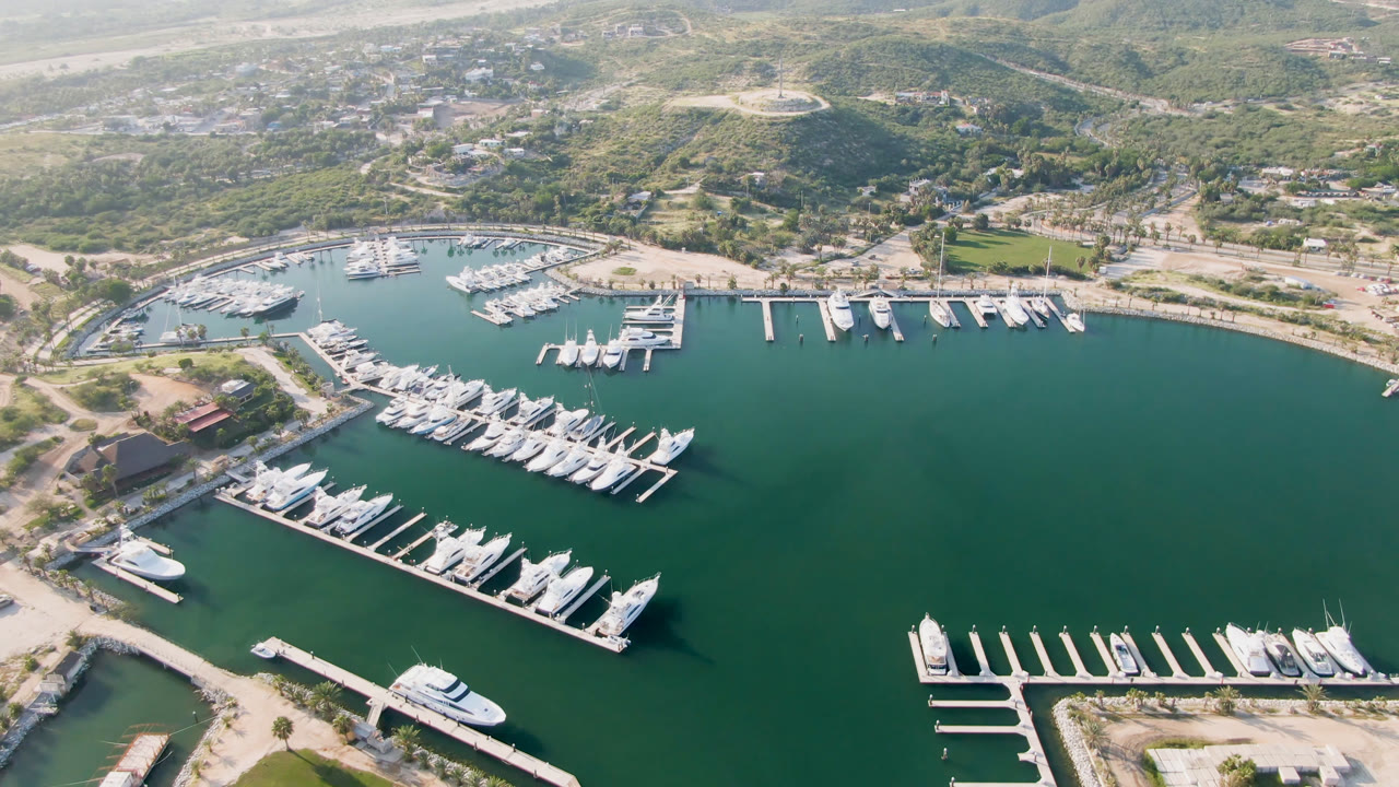 One second into the Puerto Los Cabos aerial, the marina remains centered with a long dock of yachts in the lower left, smaller slips across the water, hillside buildings, and no visible people.