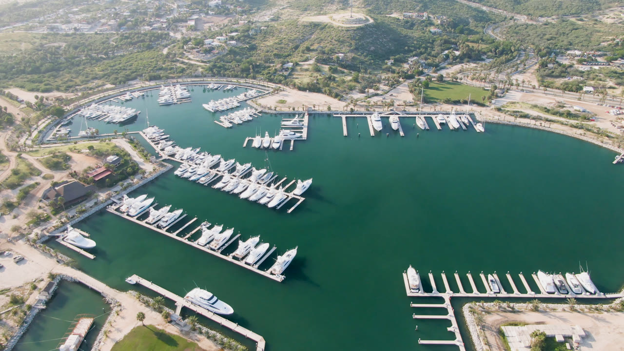 Aerial frame of Puerto Los Cabos marina with white boats docked along finger piers, green water, sandy lots, roads, and hills in the background, with no visible people.