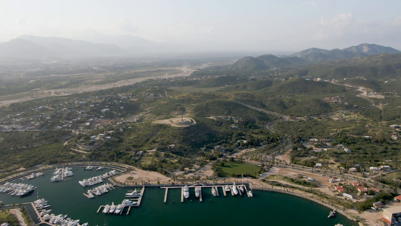 Wide aerial frame over Puerto Los Cabos with docked boats along the marina foreground, roads and low buildings beyond, hazy mountains in back, and no visible people.