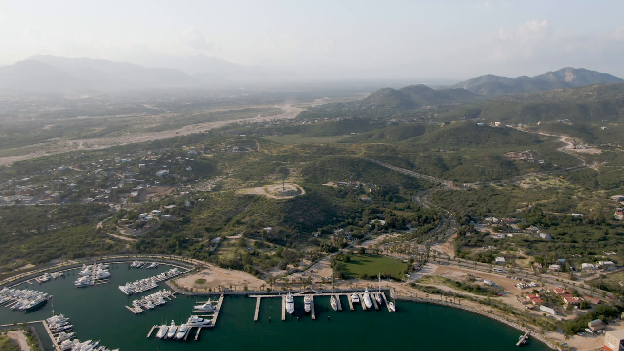 Puerto Los Cabos aerial view with white boats lined in slips at the bottom edge, a circular road feature on the hill, scattered buildings, and no visible people.