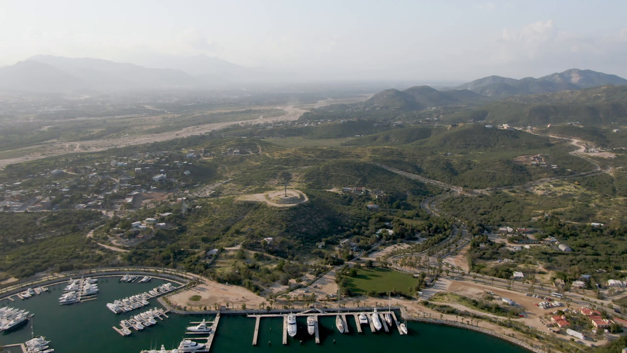 Aerial view of Puerto Los Cabos with white boats docked in green marina water, roads and low buildings inland, scrub hills, hazy mountains, and no visible people.