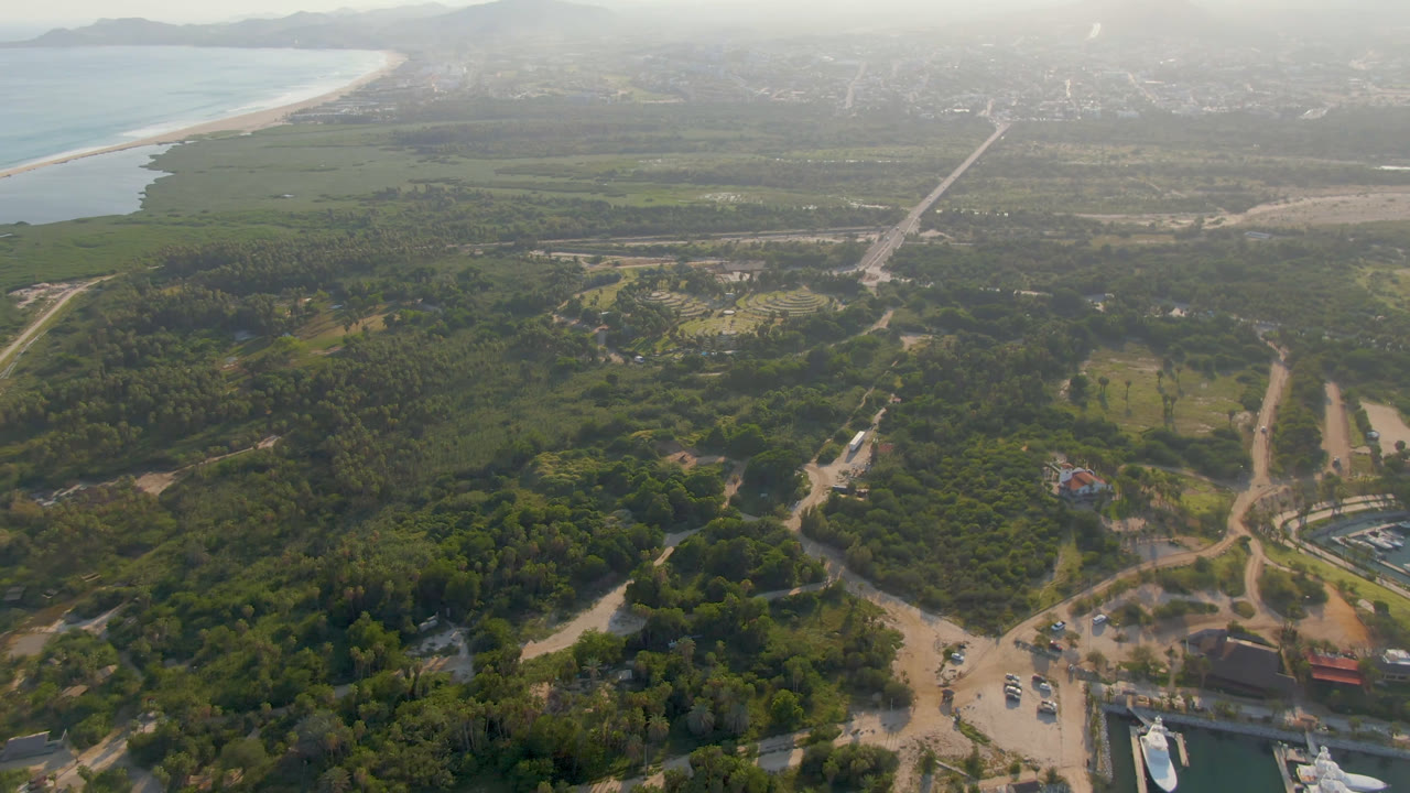 Opening aerial frame over Puerto Los Cabos with dense green vegetation, dirt roads, a sandy beach along blue ocean water, hazy distant hills, and no visible people.
