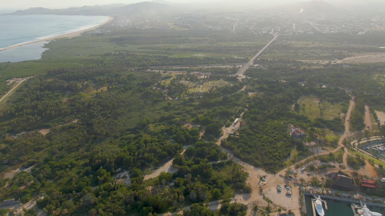 Puerto Los Cabos aerial view one second in, showing a road corridor through green land toward the hazy city edge, with the beach and ocean on the left and no visible people.