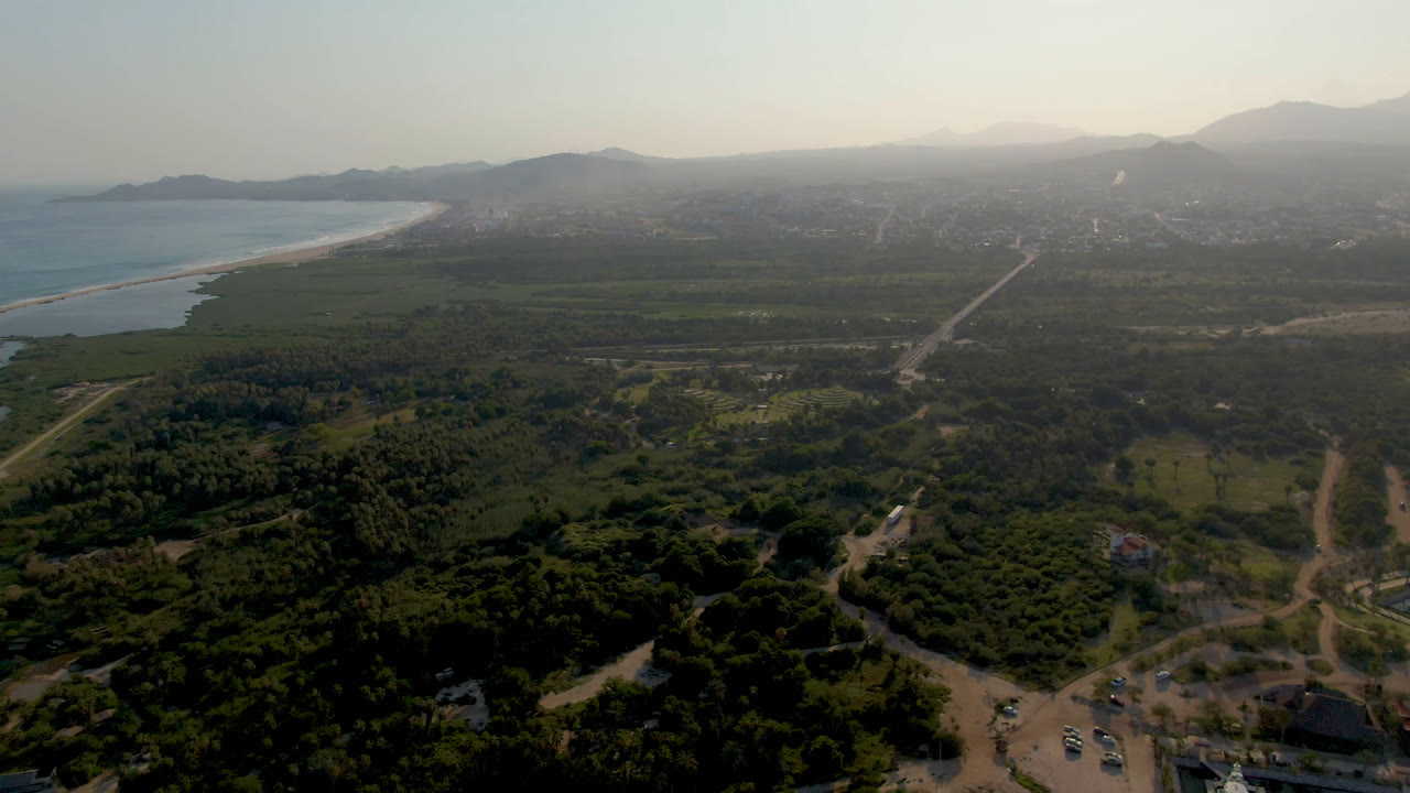 Aerial view over Puerto Los Cabos with the beach curving along the left edge, a road grid and green land below, distant development, mountains, and no visible people.