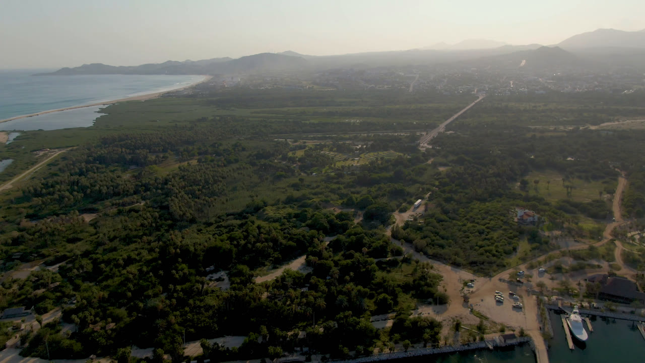 Aerial view in Puerto Los Cabos showing a boat near the marina dock at the lower edge, branching roads through green terrain, ocean to the left, distant mountains, and no visible people.