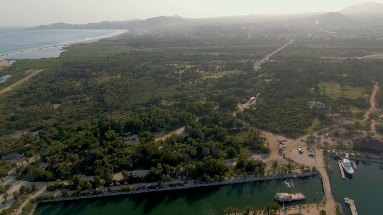 High aerial frame over Puerto Los Cabos with green marina water at the bottom, docks and boats near sandy roads, dense trees beyond, distant surf line, hazy hills, and no visible people.
