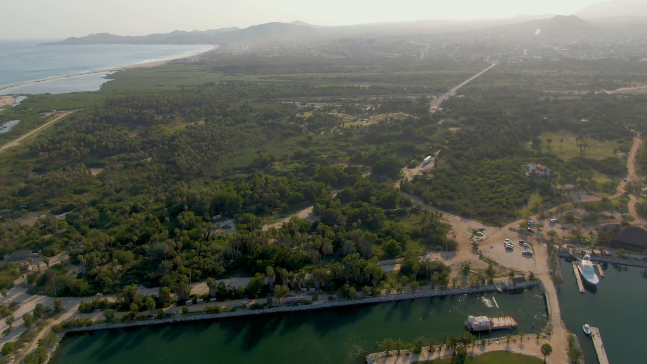 Wide Puerto Los Cabos aerial with the marina edge across the lower frame, a cluster of boats by the road, green vegetation inland, pale surf at left, hazy mountain background, and no visible people.