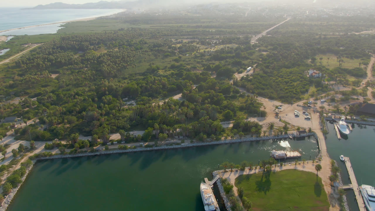 High aerial frame over Puerto Los Cabos with green marina water in the foreground, a bridge at right, white boats, tree cover, sandy roads, distant surf, and no visible people.