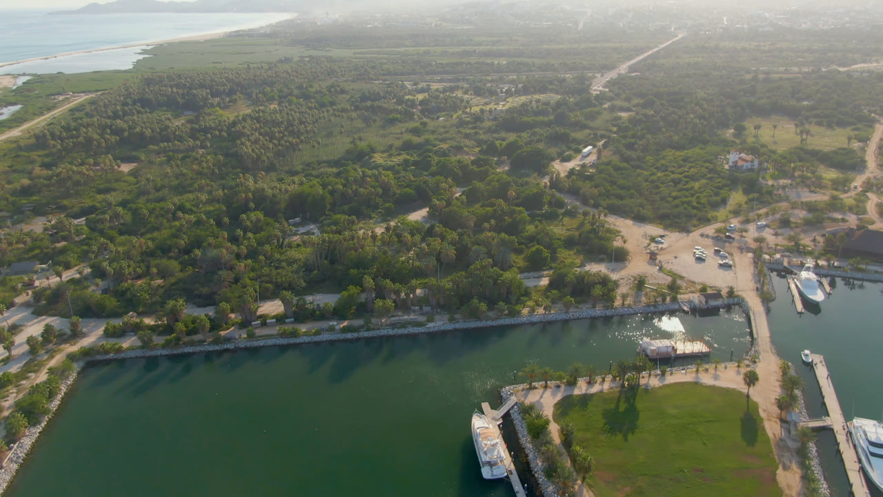 Puerto Los Cabos aerial view with the channel and bridge shifted slightly across the frame, boats near the dock, broad green vegetation, pale roads, distant ocean edge, and no visible people.