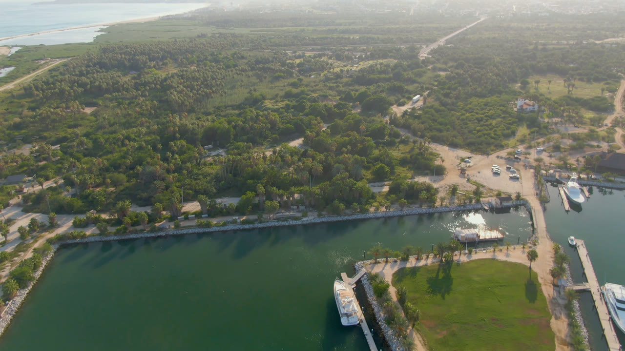 Wide aerial of a waterfront bridge, marina basin, boats, trees, and distant beach line.
