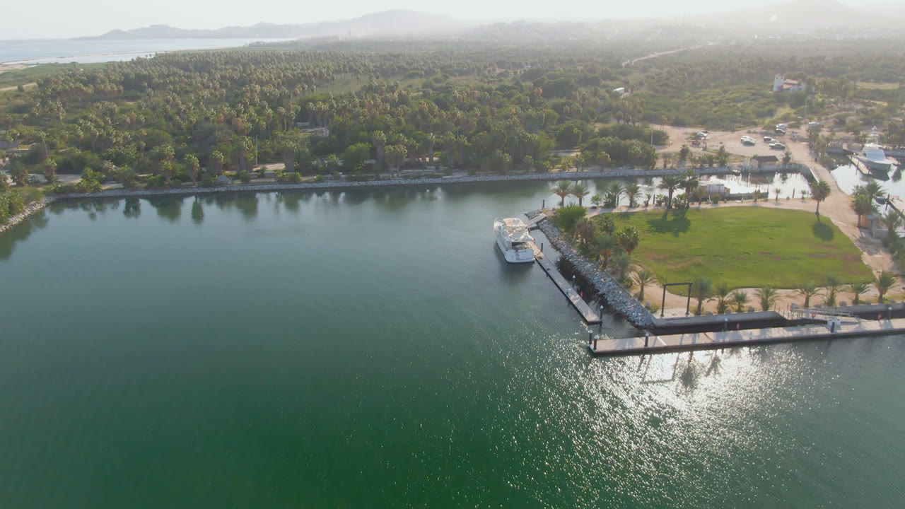 Opening aerial frame over Puerto Los Cabos water with a dock and small boat near the shore, palms and roads beyond, hazy hills in the distance, and no visible people.