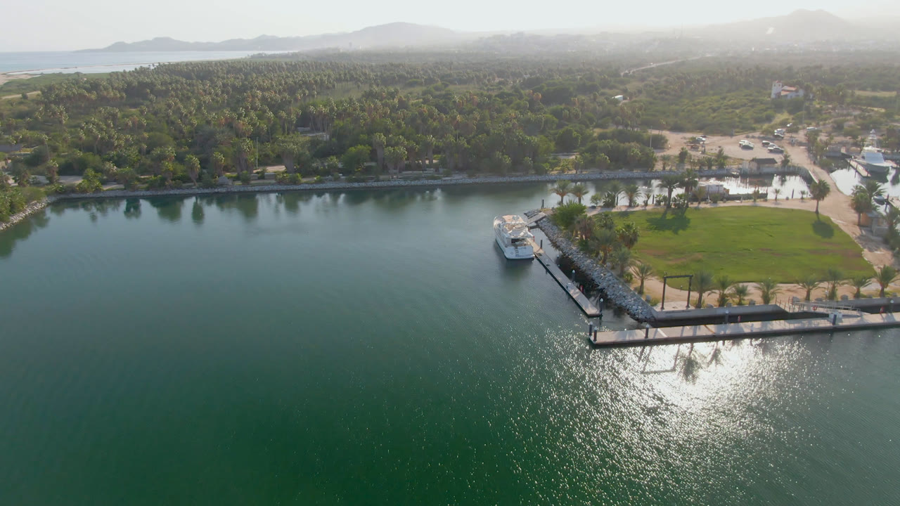 Puerto Los Cabos aerial frame with broad water in front, a dock and small boat set slightly left of center, a palm-lined shore, distant hills, and no visible people.