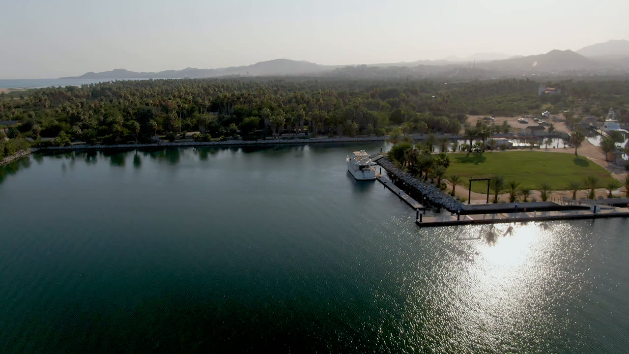 Aerial frame from Puerto Los Cabos showing green-blue water with sun reflection, a pier with a white boat near shore, palm-lined land, hazy hills, and no visible people.