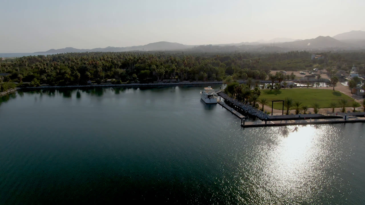 Aerial shoreline scene with reflective water, pier, boat, palms, and mountains in haze.