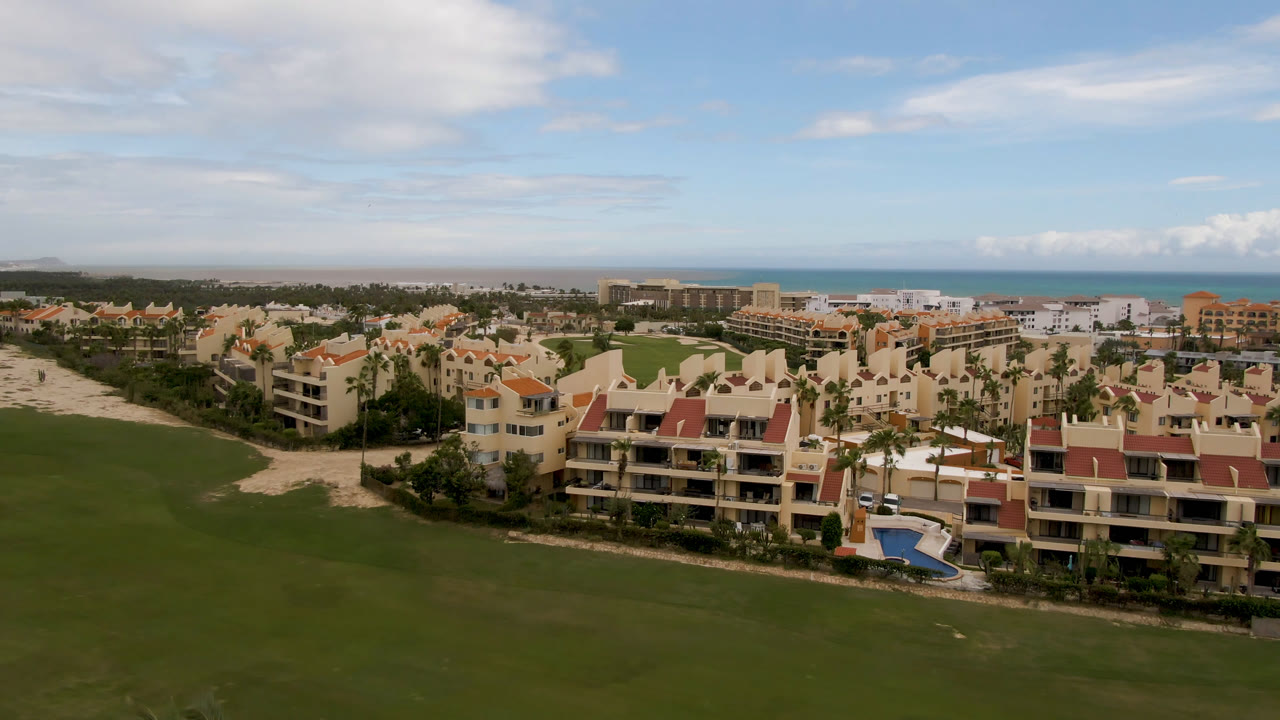 High aerial view in San Jose del Cabo showing cream resort buildings, green fairway, turquoise ocean on the horizon, scattered clouds, and no visible people.