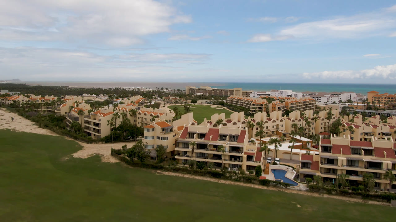 San Jose del Cabo drone wide frame with the fairway stretching across the foreground, resort blocks and palms in the midground, ocean beyond, and no visible people.