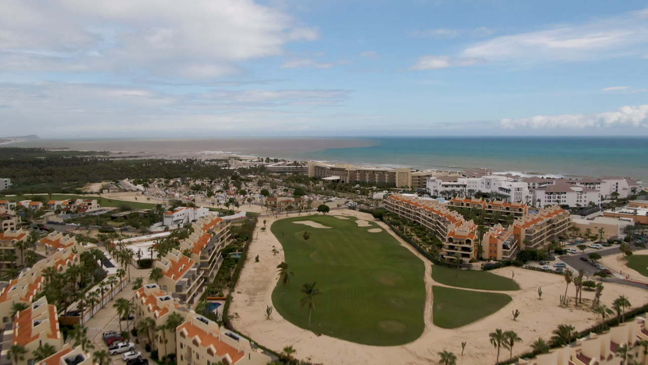 San Jose del Cabo aerial view with the fairway centered lower in frame, resort blocks along both sides, sandy shoreline beyond, blue ocean under clouds, and no visible people.
