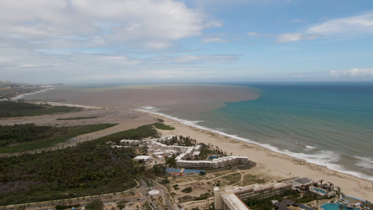 Aerial frame over San Jose del Cabo showing a resort beside a broad sandy beach, white surf lines, mixed turquoise and brown ocean water, green coastal vegetation, and no visible people.