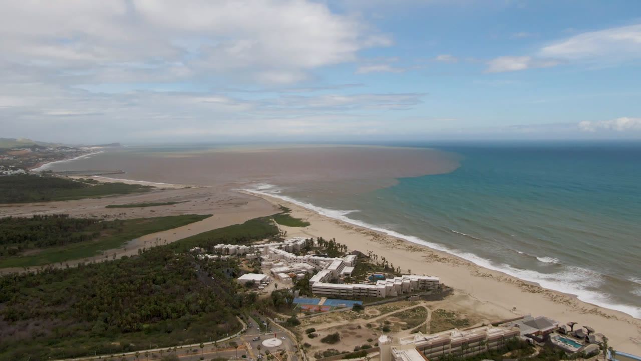 Aerial view in San Jose del Cabo with the resort lower in frame, a long beach running diagonally, surf bands along the shore, darker vegetation inland, and no visible people.