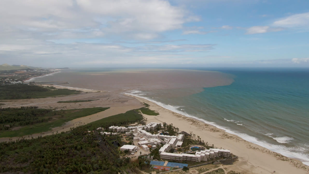 Wide aerial frame from San Jose del Cabo showing sandy beach, low surf, blue ocean beside a brown coastal water plume, green vegetation, resort buildings in the foreground, and no visible people.