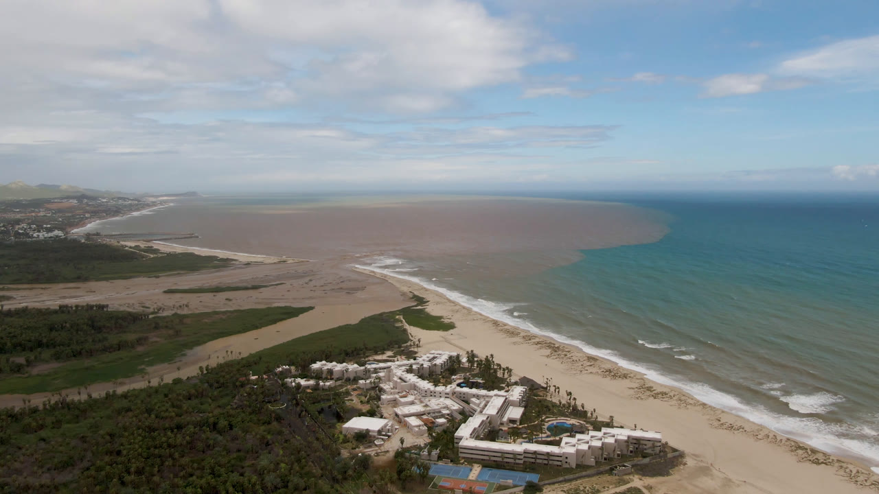 Aerial view over San Jose del Cabo coastline with surf tracing the sandy beach, turquoise ocean on the right, brown water across the bay, coastal vegetation below, and no visible people.