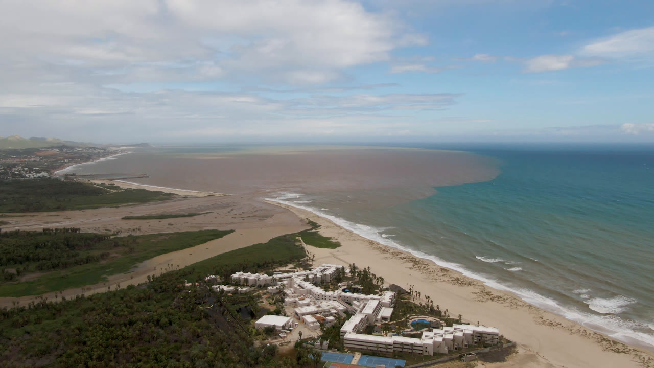 High coastal aerial showing beach, waves, brown water plume, blue ocean, resort buildings, and sky.