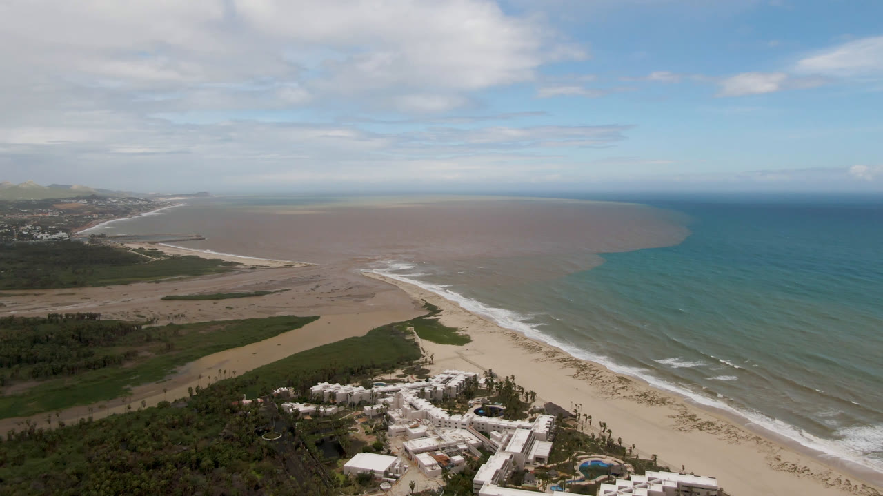 Aerial view over San Jose Del Cabo with resort buildings and palms at the bottom, a long sandy beach, white surf, blue-green ocean, brownish offshore water, scattered clouds, and no visible people.