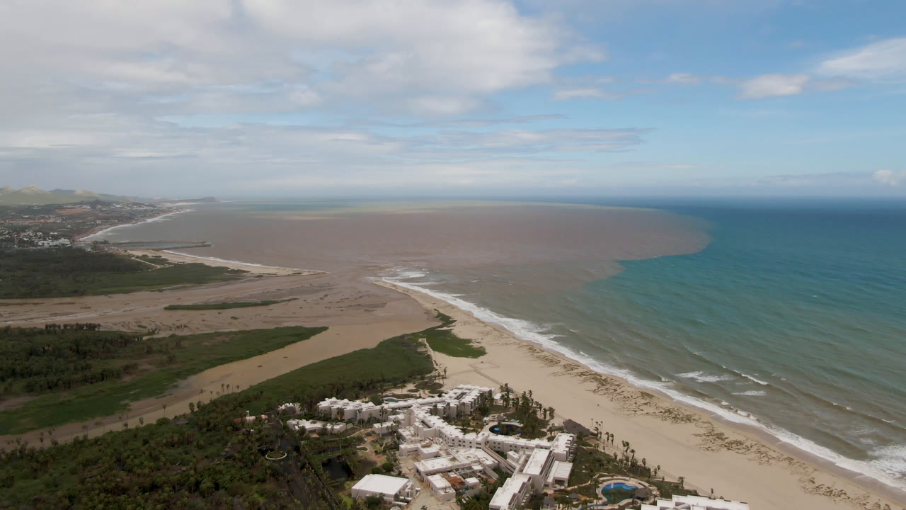 San Jose Del Cabo aerial frame with white resort roofs in the lower foreground, green coastal vegetation, a pale beach, parallel surf lines, a large brownish water area, blue ocean, and no visible people.