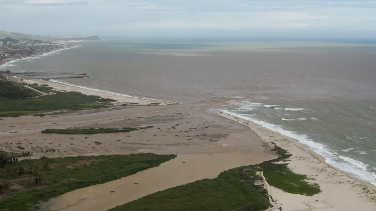 Aerial frame over San Jose del Cabo sand banks where green vegetation meets brown delta water, pale surf, and gray-blue ocean, with no visible people.