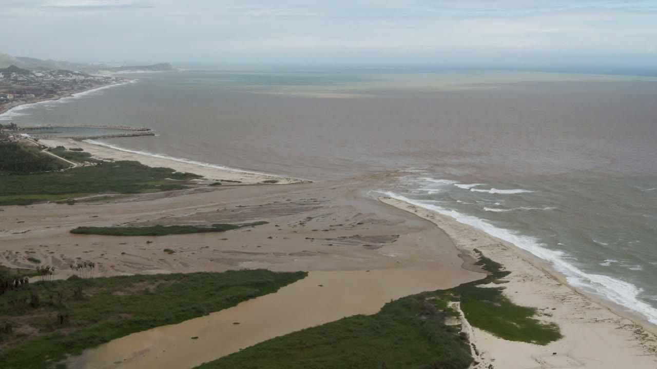 Drone-like beach view with sand flats, white waves, ocean, and distant headland.