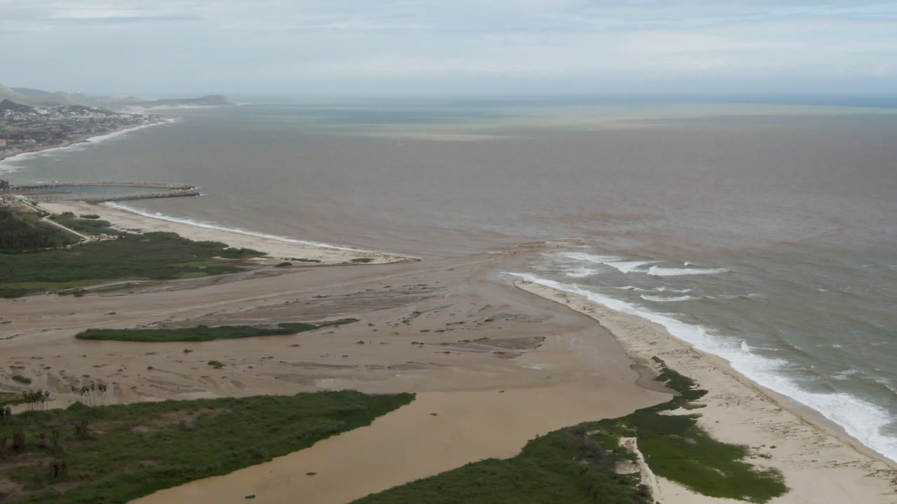 Aerial view of sand banks, surf, green vegetation, and open coastal water.