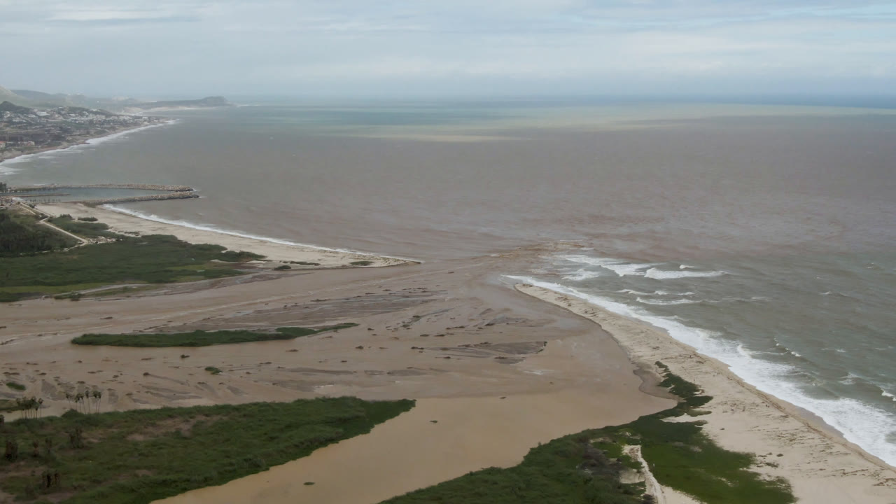 San Jose del Cabo aerial frame showing tan sand flats below, white waves along the beach, a small jetty or pier on the left coast, and no visible people.