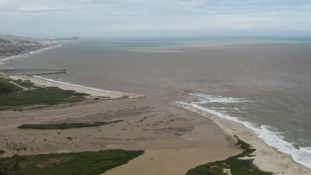 A high aerial frame over San Jose del Cabo shows open sand, green coastal vegetation, white surf lines, a jetty at left, brown-blue ocean water, and no visible people.