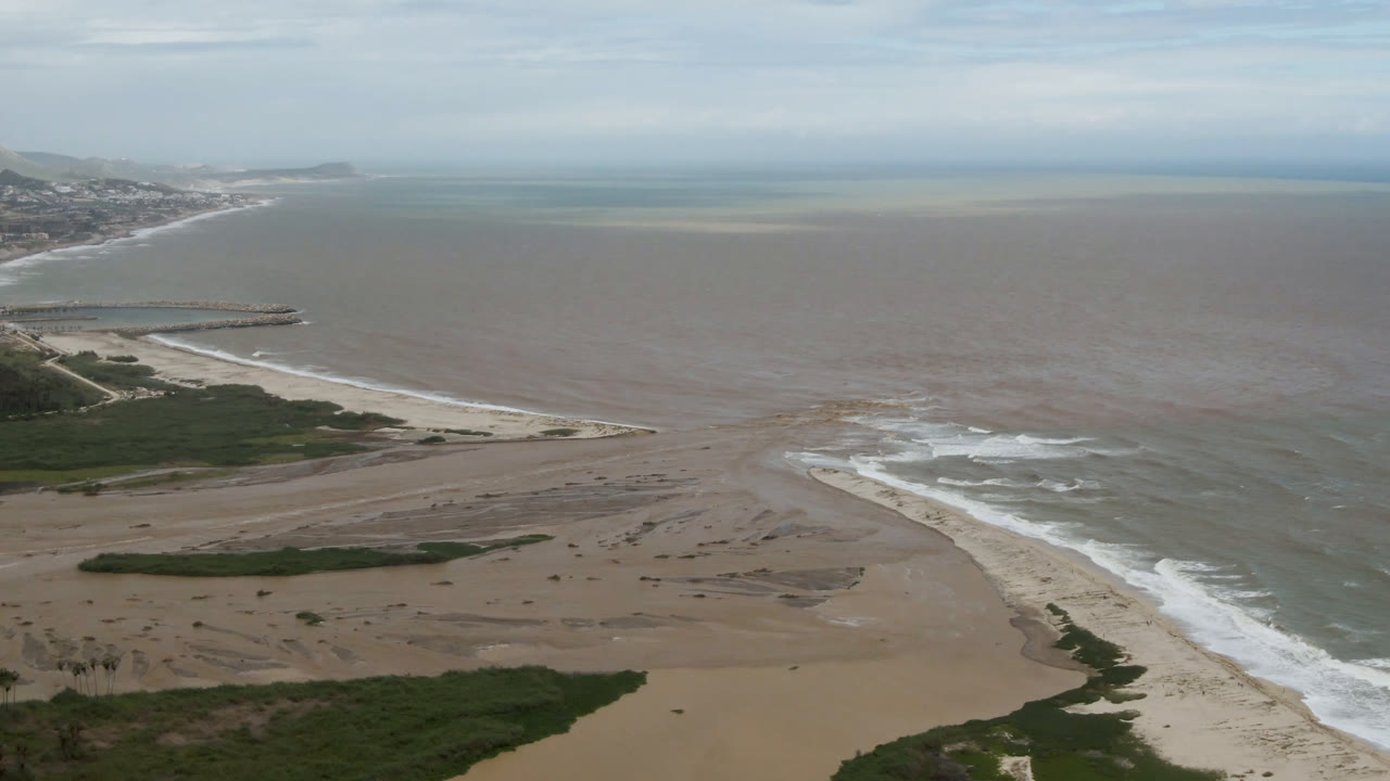 At 0:01, the San Jose del Cabo aerial view keeps the jetty and distant buildings at left while surf curves along the sand flats and no visible people appear.