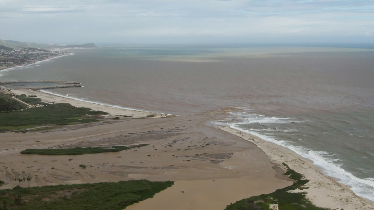Aerial beach scene with breaking waves, shallow water bands, and a long jetty.